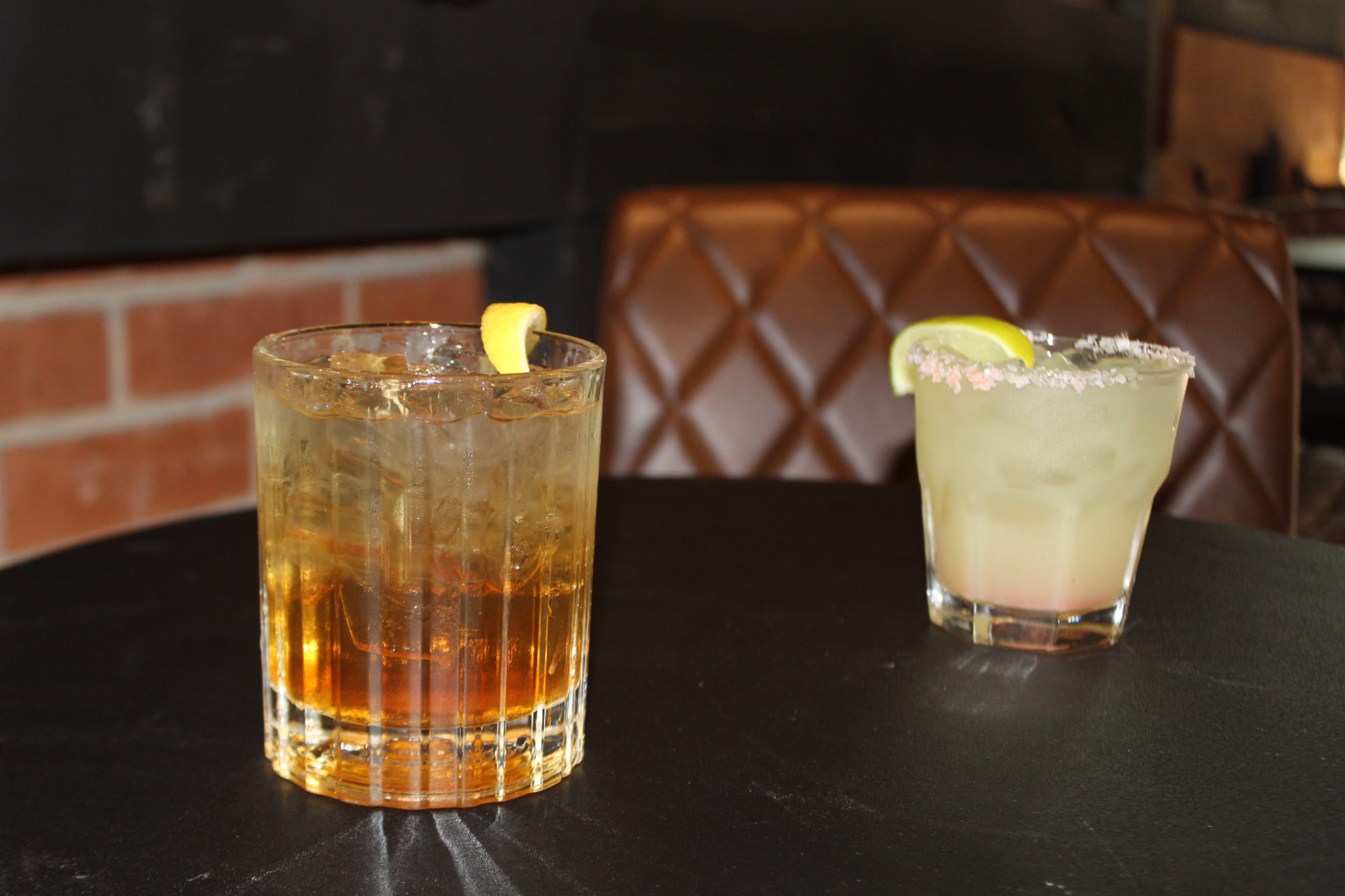 Two finished craft cocktails on a dark bar table with leather seating in the background