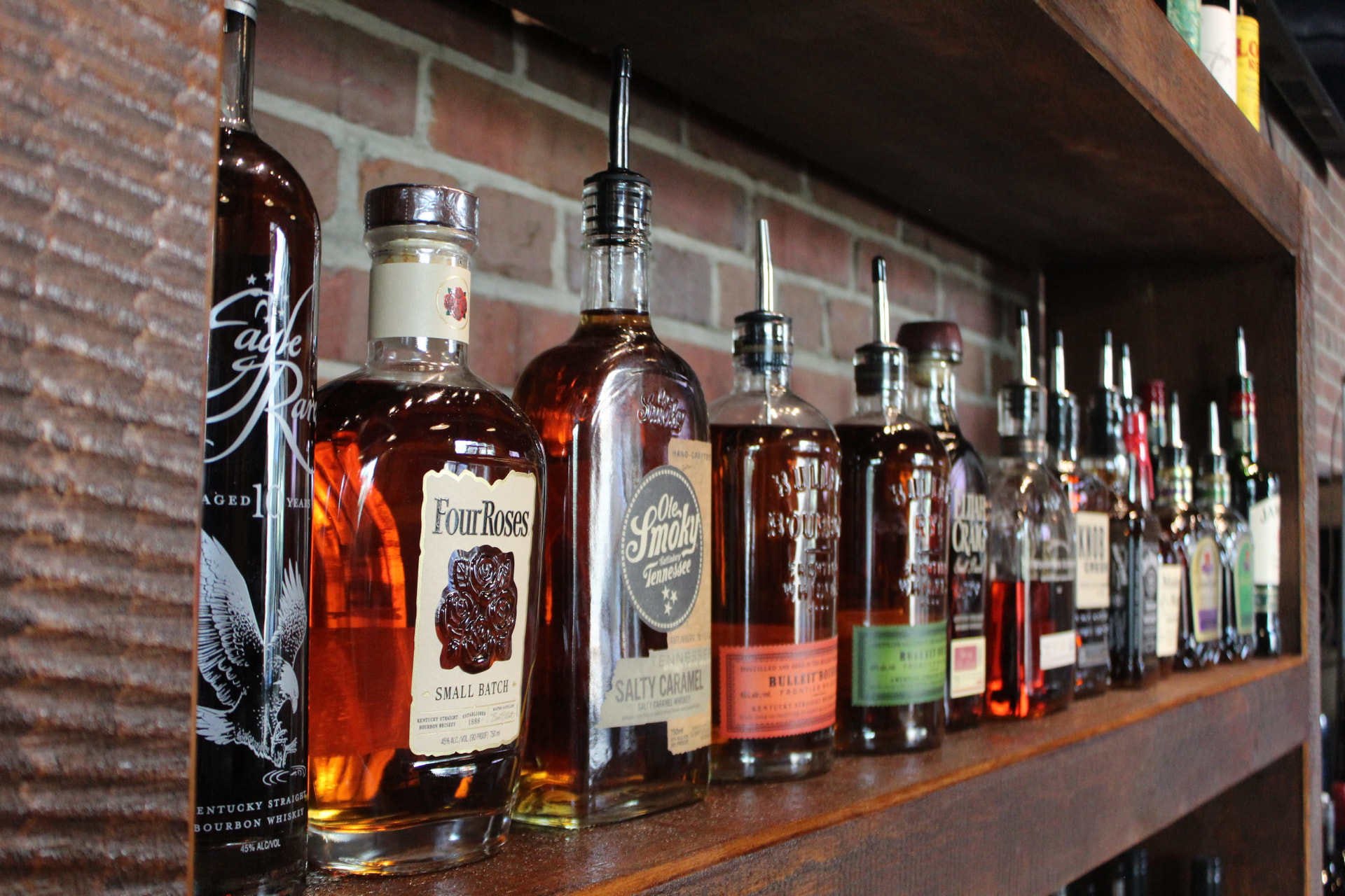 Bourbon and whiskey bottle row on a dark wood shelf against exposed brick